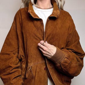 Könnte beinhalten: Braune Wildleder-Bomberjacke mit Knopfleiste, gerippten Bündchen und Kragen. Die Jacke hat zwei Vordertaschen und eine lockere Passform. Das Model trägt ein weißes Hemd darunter. Die Jacke hat einen warmen, erdigen Farbton.