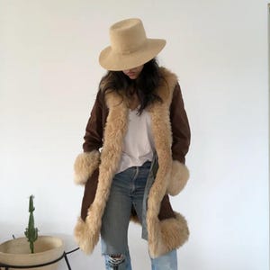 May include: A brown suede coat with tan faux fur trim and cuffs. The coat is worn with a beige hat and blue jeans. The coat has a long, flowing design, perfect for a vintage or bohemian style.
