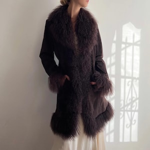 May include: A dark brown coat featuring a faux fur collar and trim. The coat has a long, flowing design with a tailored fit. The faux fur trim is a darker shade of brown, creating a contrast. The coat has pockets and is worn over a cream-coloured dress.