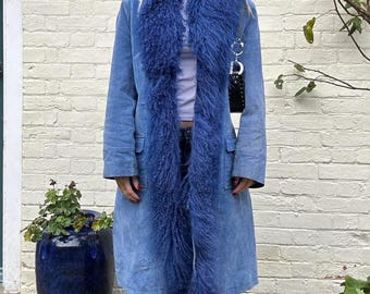 May include: A long, light blue coat with a plush, dark blue fur collar and trim. The coat has a classic design with a button closure and pockets. The coat is worn with blue jeans. A black purse hangs from the shoulder.