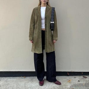 Puede incluir: Un abrigo de ante verde oliva con cuello de solapa y tres botones frontales, usado sobre un top corto blanco y pantalones negros de pierna ancha. Un bolso de hombro de cuero negro con una hebilla plateada y el texto Dior se lleva al hombro.