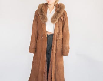 May include: A long, brown suede coat with a faux fur collar. The coat is double-breasted and reaches the ankles. The model is wearing a white shirt and dark pants. The coat has a vintage aesthetic.