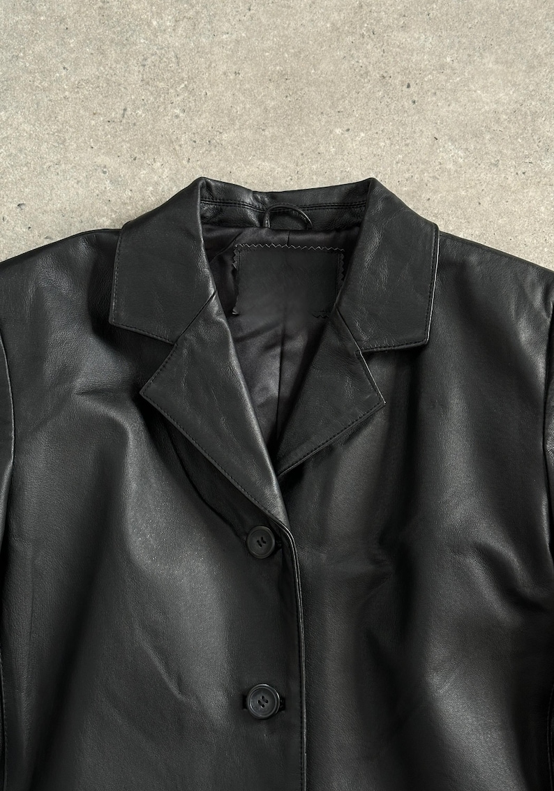 May include: A black leather blazer with a notched lapel collar and two button closure. The jacket has a classic design and is made of a smooth, shiny material. The blazer is a versatile piece of outerwear.