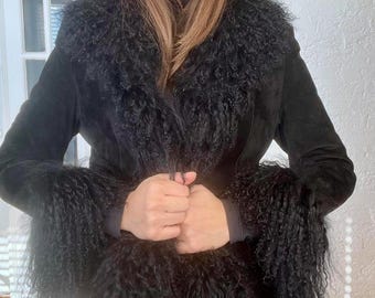 Puede incluir: Una chaqueta de ante negra con un gran cuello de piel negra esponjosa y puños. La chaqueta está ceñida a la cintura y tiene un estilo largo y fluido.