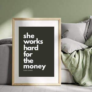 Könnte beinhalten: Ein gerahmter Druck mit dem weißen Text "she works hard for the money" auf dunklem Hintergrund. Der Druck befindet sich in einem hellen Holzrahmen. Das Zitat wird Donna Summer zugeschrieben.