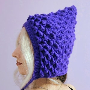 May include: A striking purple crochet hat with a pointed crown and textured pattern. The hat has ear flaps and is crafted from a thick, knitted material. This handmade hat is a unique accessory.