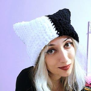Könnte beinhalten: Ein zweifarbiges Häkelhut in Schwarz und Weiß. Der Hut hat spitze Ohren, eine weiche Textur und ein einzigartiges Farbblock-Design. Der Hut ist handgefertigt.