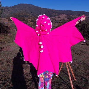 Puede incluir: Un poncho rosa brillante con capucha, adornado con pompones blancos. El poncho está extendido, mostrando su forma. La capucha presenta lunares blancos. La persona lleva leggings estampados. El fondo incluye una montaña y cielo azul.
