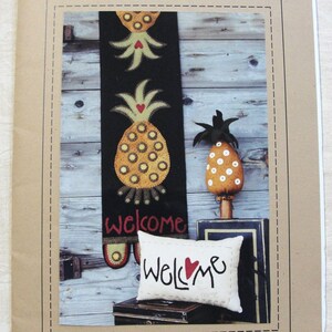 May include: A decorative pineapple-themed display featuring a black runner with pineapple designs and the word "welcome". A small pillow with "Welcome" and a heart is also present, along with a pineapple ornament.