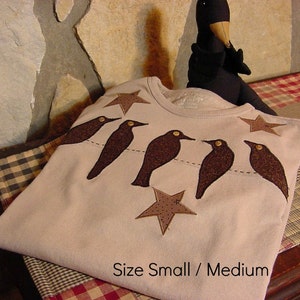 May include: A light beige long-sleeved shirt featuring appliqued brown birds perched on a dotted line, with brown star accents. The text "Size Small / Medium" is printed on the shirt.