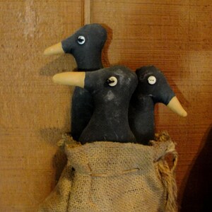 Peut inclure: Trois poupées en forme d'oiseaux gris foncé avec des becs crème et des yeux blancs, nichées dans un sac en toile de jute. Les poupées ont des détails d'yeux en bouton ou cousus. Le fond est une surface en bois.