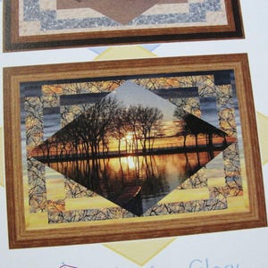 May include: Framed artwork depicting a sunset over a lake, with trees silhouetted against the golden light. The artwork is set within a diamond shape, surrounded by a patterned border and a wooden frame.