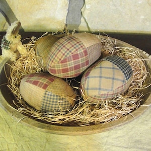 May include: A wooden bowl filled with straw and three plaid fabric Easter eggs. The eggs have patches of red, blue, and tan fabric. A small stuffed bunny with a plaid waistcoat sits on the edge of the bowl.