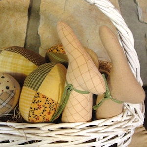 May include: A white wicker basket filled with decorative Easter eggs and bunny-shaped ornaments. The eggs are covered in patchwork fabric with plaid and floral patterns. The bunny ears are beige and tied with green ribbon.