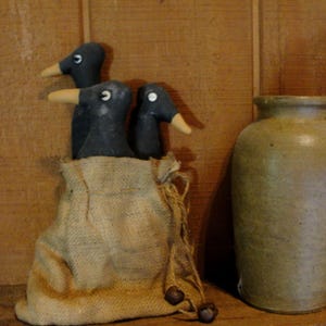 May include: Three black bird figures with yellow beaks are in a burlap sack. An olive-green ceramic jar is to the right. The background is a wooden surface, creating a rustic, country aesthetic.