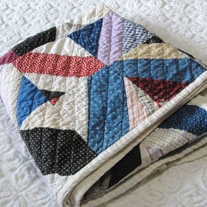 May include: A folded patchwork quilt featuring a variety of fabric patterns in shades of blue, red, black, white, and lavender. The quilt is constructed from triangular and rectangular fabric pieces with visible stitching throughout the surface.