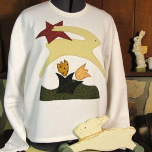 May include: A white sweatshirt with long sleeves featuring a whimsical bunny and star applique design. The bunny is yellow and the star is burgundy. The design includes green and orange floral accents, creating a spring or Easter theme.