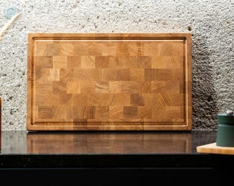 May include: A rectangular wooden chopping board with a juice groove. The board is made of end-grain wood, with a light brown colour and a checkered pattern. The board is on a black surface.
