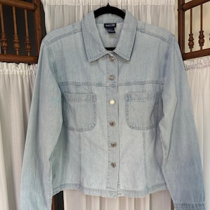May include: Light blue denim jacket with a collared neckline, two chest pockets, and button closures. The jacket has long sleeves and a classic design. The brand name "BACCOM" is visible on the label.