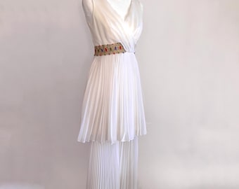 May include: A white pleated dress with a V-neck bodice and a decorative belt. The belt is adorned with colorful beads. The dress is displayed on a white mannequin against a white background.