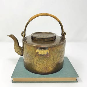 May include: Antique brass teapot with a dragon-shaped spout and a woven handle. The teapot features intricate embossed dragon scale patterns and sits on a teal book. A small gold-colored plaque is on the front of the teapot.