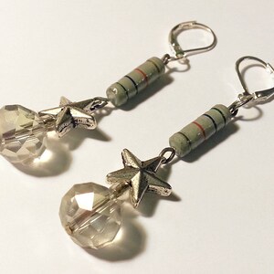 May include: A pair of dangle earrings featuring silver-toned leverback hooks, a cylindrical light green component with thin blue and red stripes, a five-pointed silver-toned star charm, and a clear, multifaceted spherical glass bead at the base.