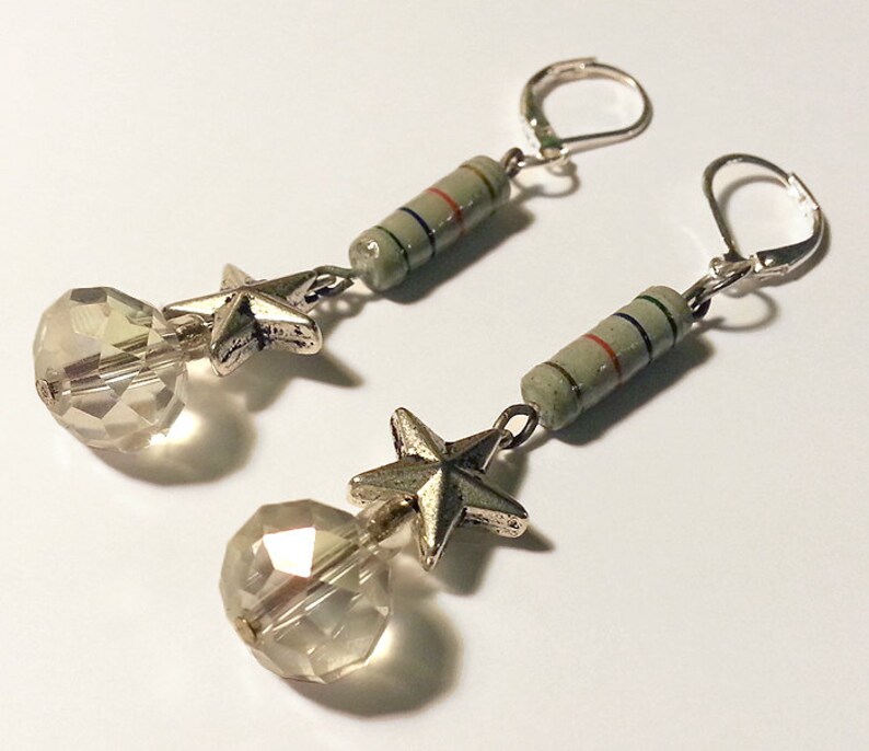 May include: A pair of dangle earrings featuring silver-toned leverback hooks, a cylindrical light green component with thin blue and red stripes, a five-pointed silver-toned star charm, and a clear, multifaceted spherical glass bead at the base.