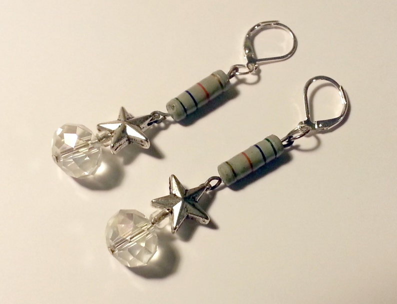 May include: A pair of dangle earrings featuring silver-toned leverback hooks, a cylindrical gray electronic resistor component with thin colored stripes, a five-pointed silver-toned star charm, and a clear faceted spherical glass bead at the base.