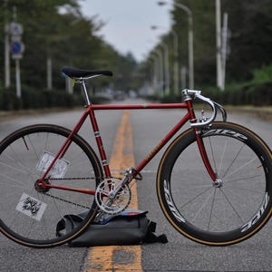 Könnte beinhalten: Ein metallisch rotes Rennrad mit einem dünnen Stahlrahmen, Rennlenker und schwarzen Zipp-Laufrädern. Das Fahrrad verfügt über eine silberne Kurbelgarnitur, einen schwarzen Sattel und ein kleines weißes rechteckiges Schild an den Speichen.