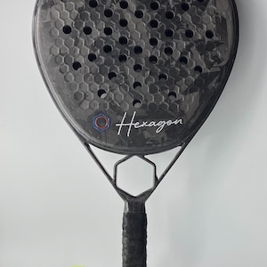 Peut inclure: Une raquette de paddle-tennis noire et grise avec un motif hexagonal et le mot "Hexagon" imprimé sur la face. Une balle de tennis jaune est attachée à la raquette avec une sangle noire.