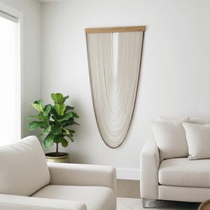 Puede incluir: Un moderno tapiz de pared con una parte superior de madera clara y hebras fluidas de color blanquecino. La obra de arte está montada en una pared blanca, complementando los tonos neutros de la habitación. Una planta en maceta verde y muebles color crema completan el diseño.