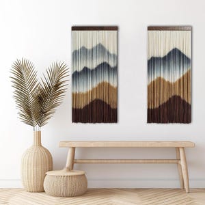 May include: Two mountain-themed wall hangings with a gradient color scheme. The top is a light cream color, transitioning to blue, then brown, and finally a dark brown at the bottom. The hangings are mounted on a dark brown wooden bar.
