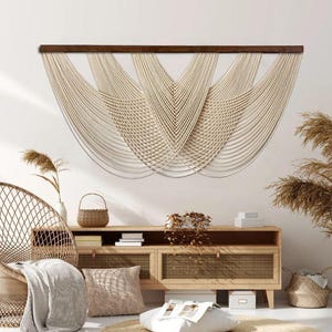 May include: A large, cream-colored macrame wall hanging with a dark brown wooden bar. The design features multiple cascading, curved lines, creating a layered, textured effect. The wall hanging is a decorative element for a living room.