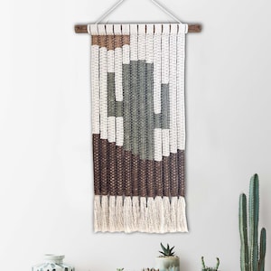 Op de afbeelding: Een geweven wandkleed met een cactusontwerp. De wandtapijt heeft een cactusvorm in tinten groen en wit, met bruine en witte geweven secties. De onderkant heeft franjes. De tekst "MODEL A" staat in de linkerbovenhoek.