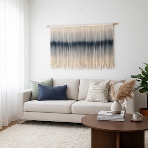Op de afbeelding: Een woonkamer met een beige bank, kussens en een ronde houten salontafel. Boven de bank hangt een wandkleed met een blauw en crème ombre-ontwerp. De kamer heeft ook een wit tapijt en een plant in een pot.