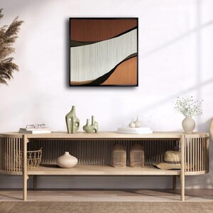 May include: A light wood console table with two shelves. The table has a curved design and is decorated with a woven basket, a vase, and a hat. A framed abstract art print with brown, white, and black colours hangs on the wall behind the table.