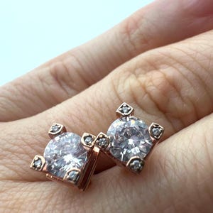 May include: A pair of rose gold stud earrings. Each earring features a large, round, clear gemstone, surrounded by smaller accent stones. The earrings have a square design with a small accent stone at each corner.