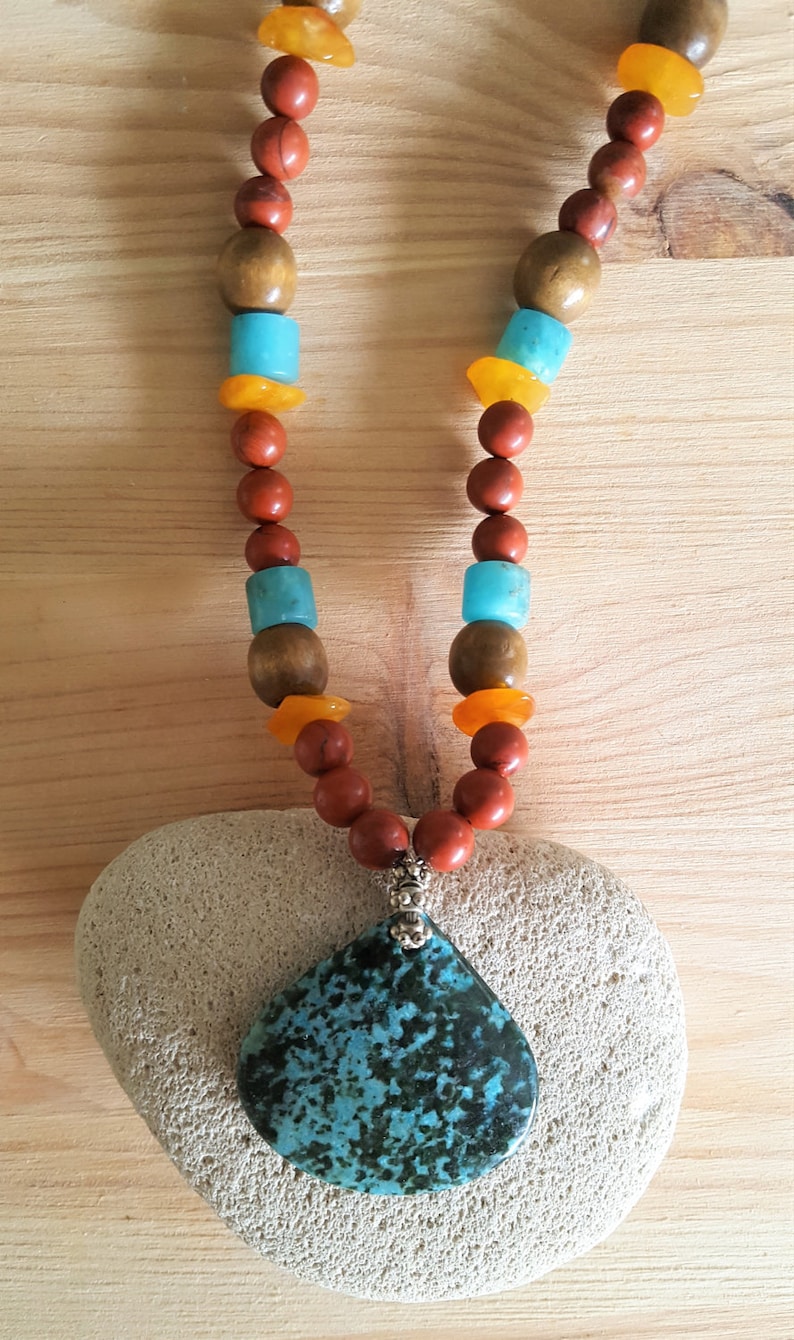 Puede incluir: Un collar con un colgante de piedra turquesa y cuentas rojas, marrones y naranjas. El collar est&aacute; sobre una piedra blanca.