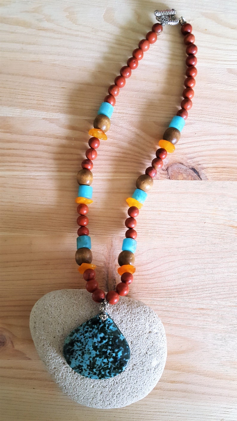 Puede incluir: Un collar hecho de cuentas marrones, naranjas y turquesas con un gran colgante de piedra turquesa. El collar est&aacute; sobre una superficie de madera marr&oacute;n claro.