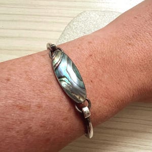 May include: Silver bracelet featuring an oval abalone shell centerpiece with iridescent blues, greens, and purples. The bracelet is attached to a silver band with a simple clasp. The jewelry is worn on a wrist.