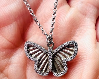 May include: A silver-colored butterfly pendant necklace. The butterfly has a detailed design with a textured body and wings outlined with small, clear stones. The necklace chain is also silver-colored.