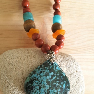 Puede incluir: Un collar con un colgante de piedra turquesa y negra, con cuentas marrones, naranjas y azules. El collar est&aacute; sobre una piedra blanca.