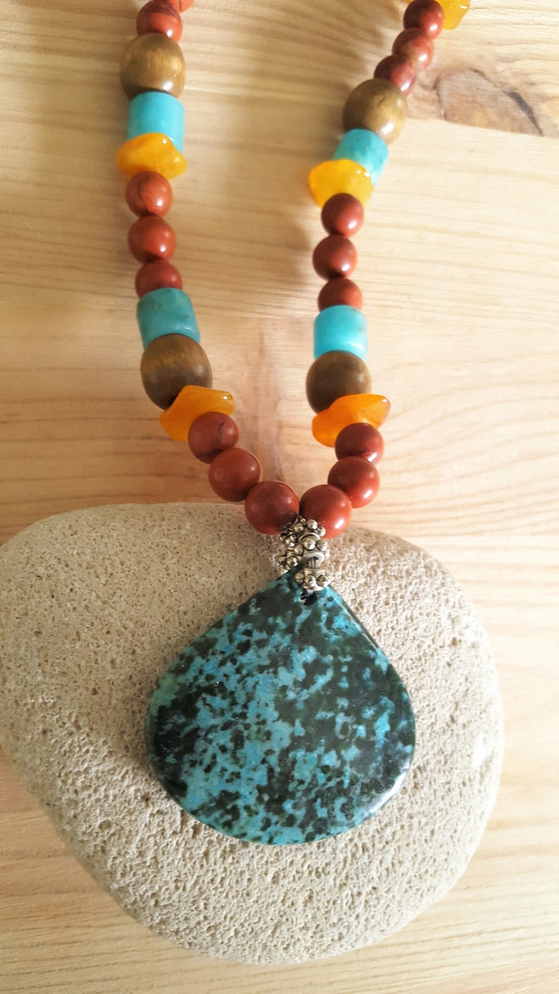 Puede incluir: Un collar con un colgante de piedra turquesa y negra, con cuentas marrones, naranjas y azules. El collar est&aacute; sobre una piedra blanca.