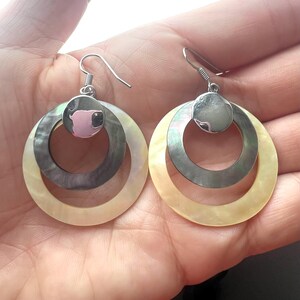 May include: A pair of silver-tone hoop earrings. Each earring features three concentric circles: a pearlescent outer ring, a dark gray middle ring, and a small silver top circle. The earrings are held by a silver fish hook.