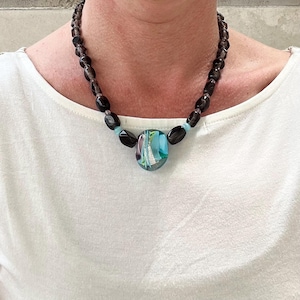 Op de afbeelding: Een zwarte kralenketting met een grote, ronde, veelkleurige glazen hanger. De hanger heeft blauwe, groene en witte swirls.