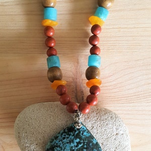 Puede incluir: Un collar con un colgante de piedra turquesa y cuentas rojas, marrones y naranjas. El collar est&aacute; sobre una piedra blanca.