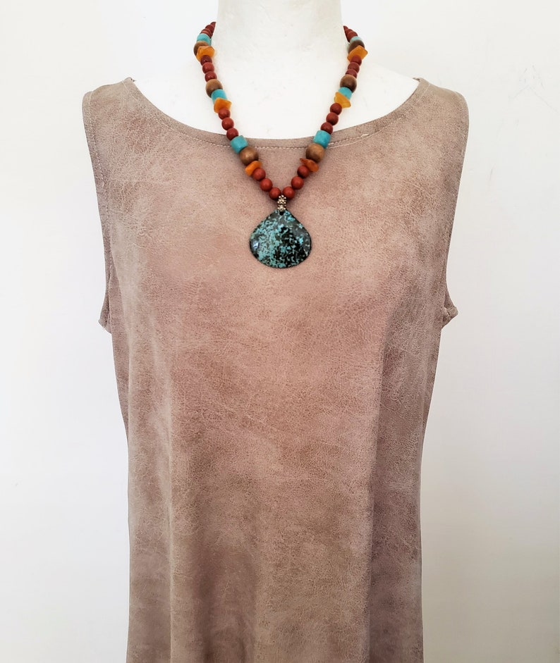 Puede incluir: Un collar con un colgante de piedra turquesa y cuentas marrones, naranjas y azules. El collar est&aacute; sobre un fondo de ante marr&oacute;n.