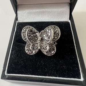 May include: A silver-toned butterfly ring with dark gray stones, presented in a black jewelry box. The butterfly design features detailed wings and body, showcasing a delicate and elegant aesthetic. The ring is a statement piece.