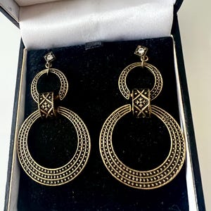 Puede incluir: Un par de pendientes de color dorado con un diseño de doble círculo. Los pendientes tienen una superficie texturizada y una pequeña piedra transparente en la parte superior.