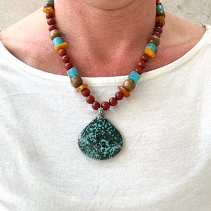 Puede incluir: Un collar de cuentas turquesa y marr&oacute;n con un gran colgante turquesa. El collar est&aacute; hecho con cuentas rojas, marrones y turquesas.